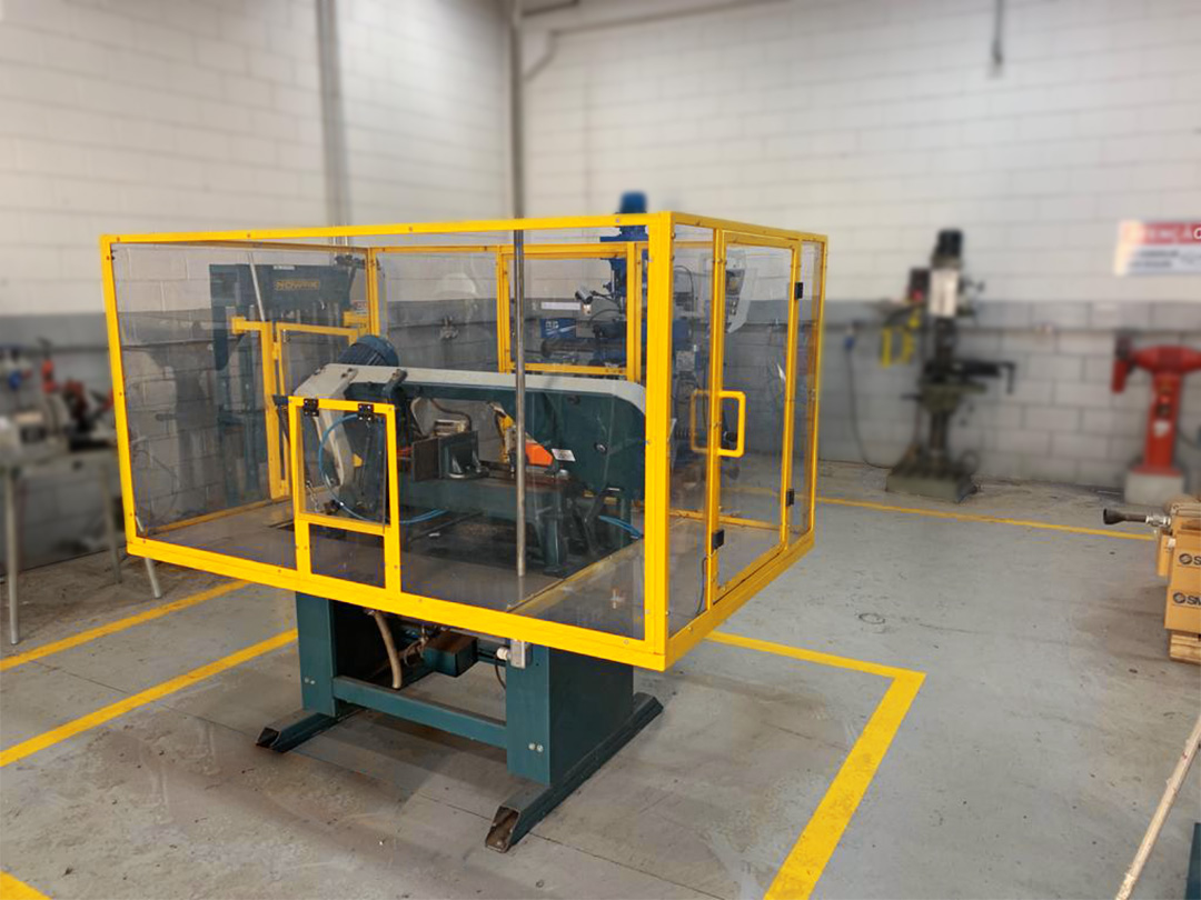 Máquina industrial de usinagem protegida por gaiola de segurança em grade amarela, em ambiente de produção com sinalização de segurança no piso.