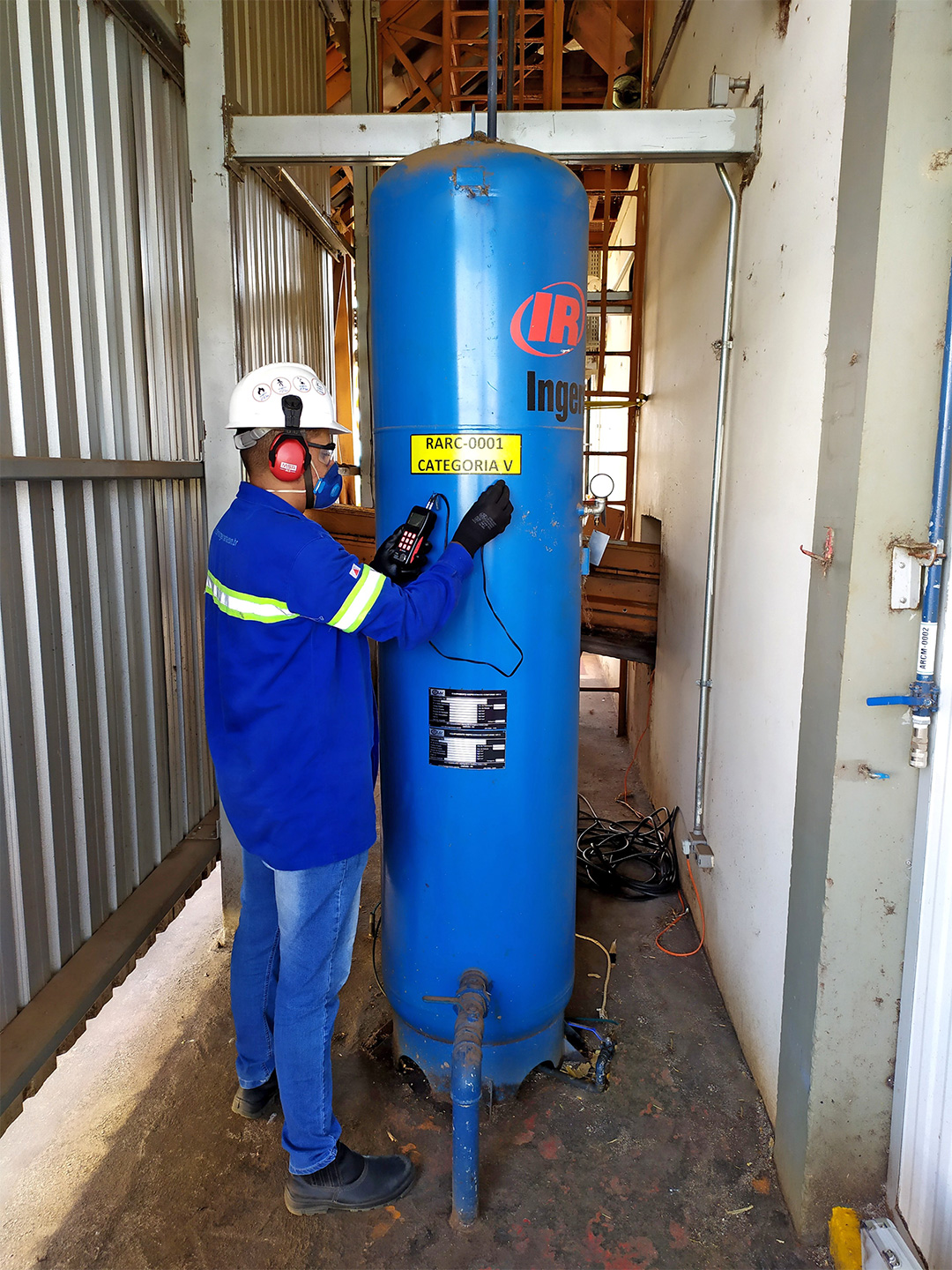Funcionário com capacete e uniforme azul realizando manutenção ou inspeção em um reservatório de ar comprimido vertical de cor azul, localizado em ambiente industrial.