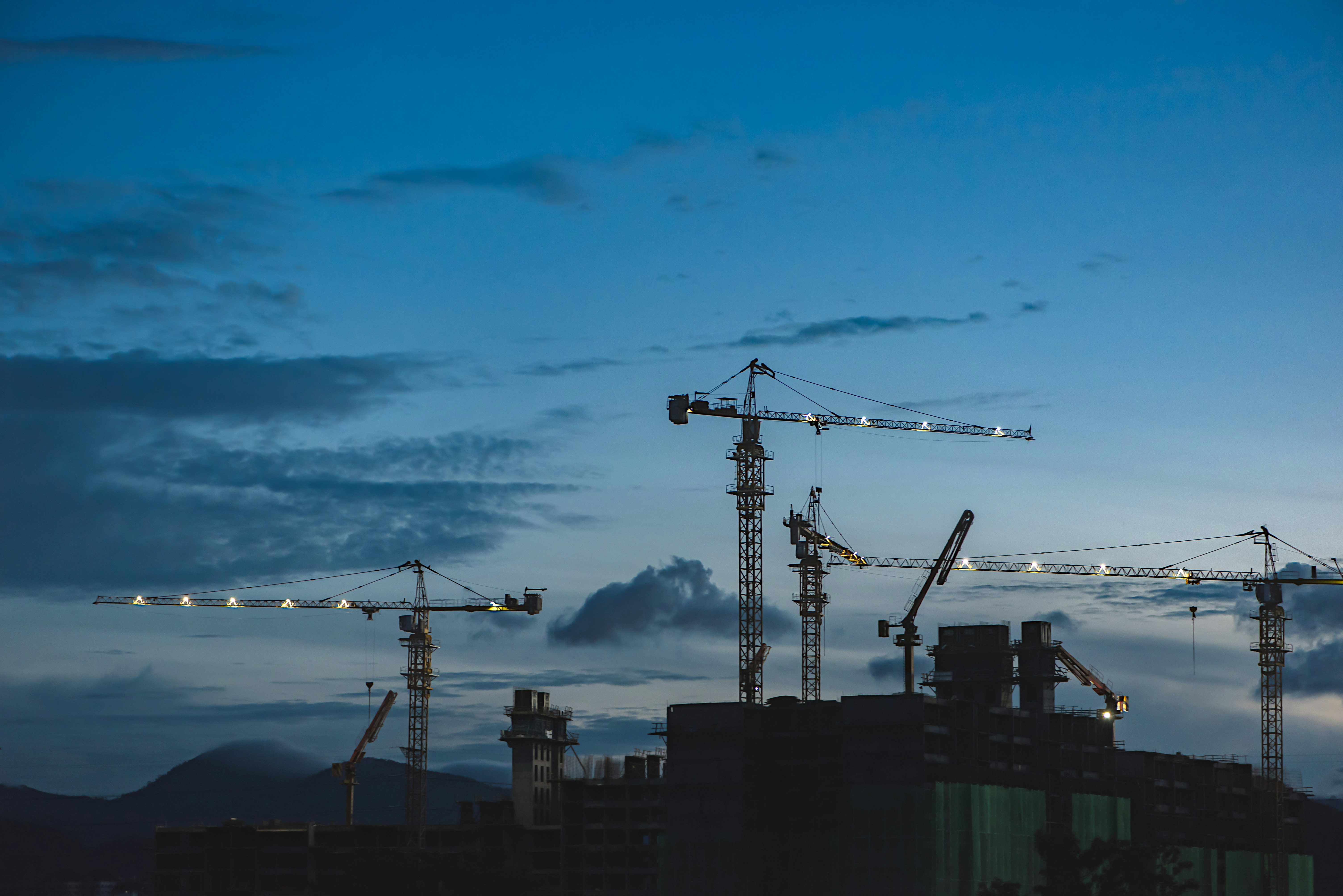 Canteiro de obras de grande porte, com estrutura metálica de galpão em construção, edifício de concreto e guindastes operando sob céu claro
