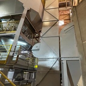 Elevador de canecas vertical com estrutura metálica autoportante instalado em planta industrial, com seção de descarga superior, base de carregamento e passarelas de manutenção com guarda-corpo amarelo.