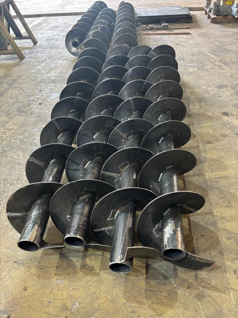 Conjunto de roscas transportadoras helicoidais em aço carbono, com eixos tubulares e lâminas soldadas, dispostas paralelamente em piso de galpão industrial.