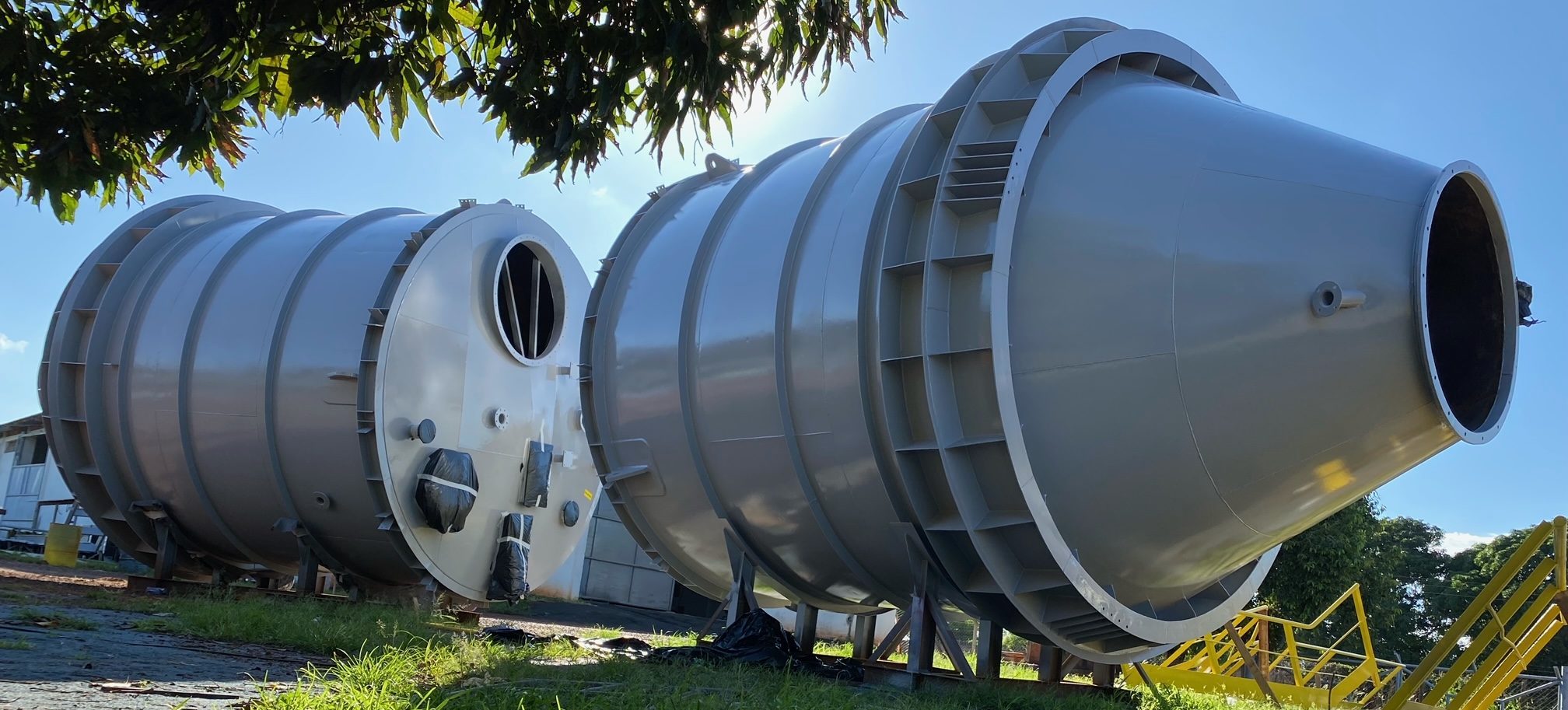 Silos metálicos de grande porte com extremidade cônica dispostos em área de armazenamento ao ar livre. Equipamentos de caldeiraria pesada representados comercialmente pela Ferrbá.